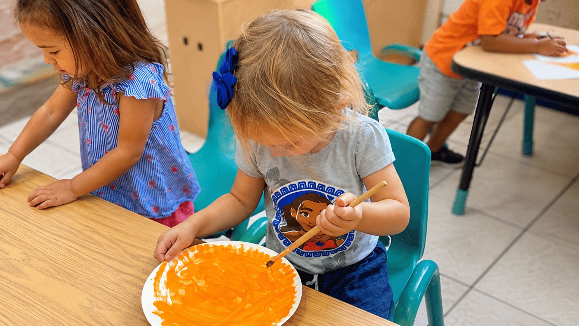 Preschooler painting a paper plate orange at a classroom table